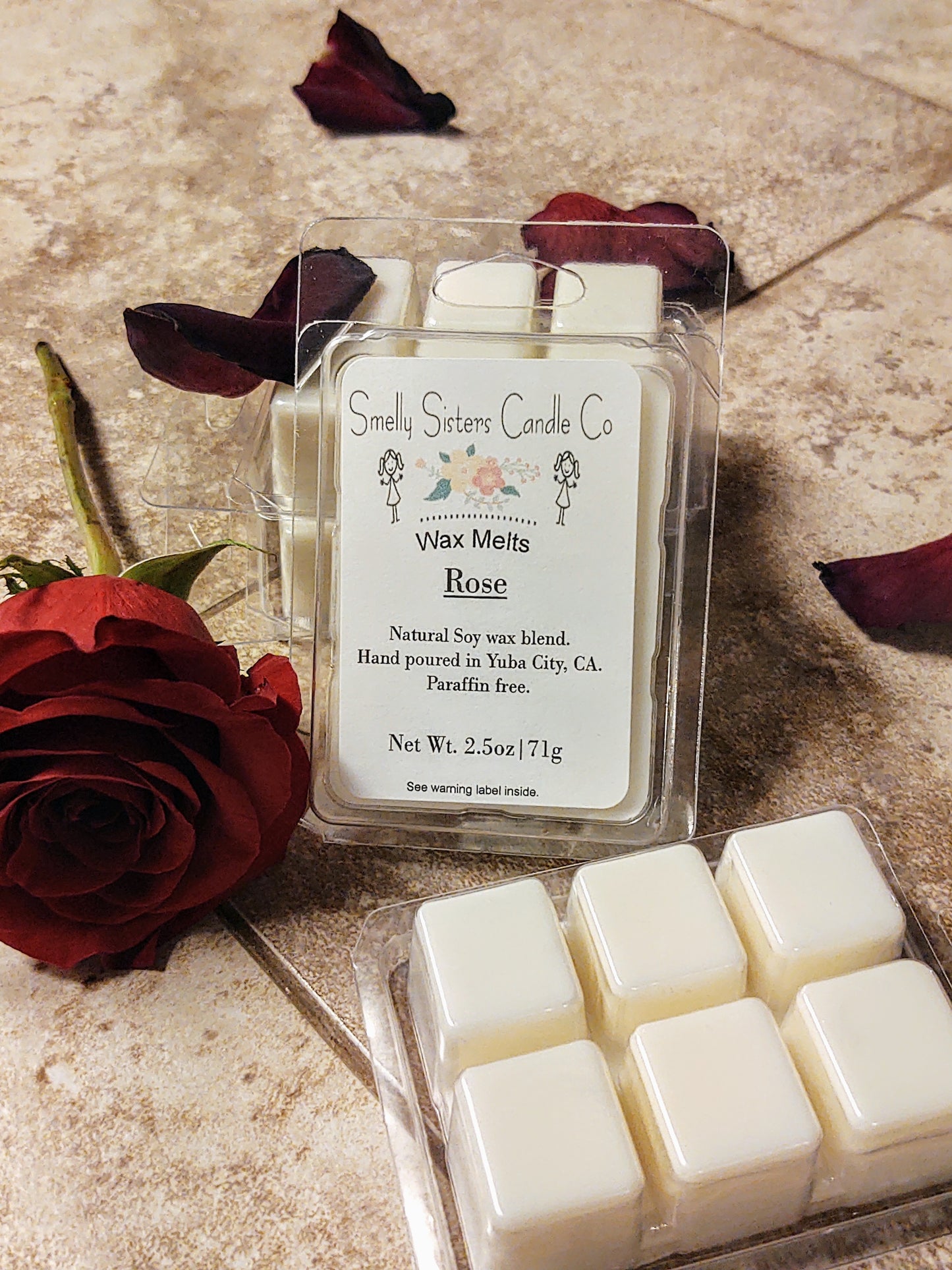 A package of wax melts with a rose scent, displayed on a surface with rose petals around it. The packaging is clear, showcasing the wax cubes inside, and there is a label with the product details.
