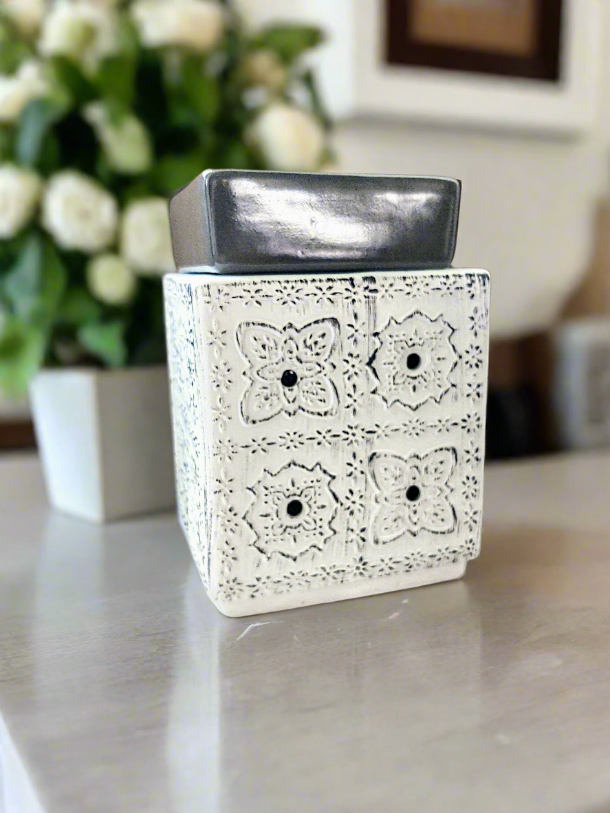 A white wax warmer with a geometric pattern and a silver top, placed on a table. Neutral. Larger size. House warming gift.