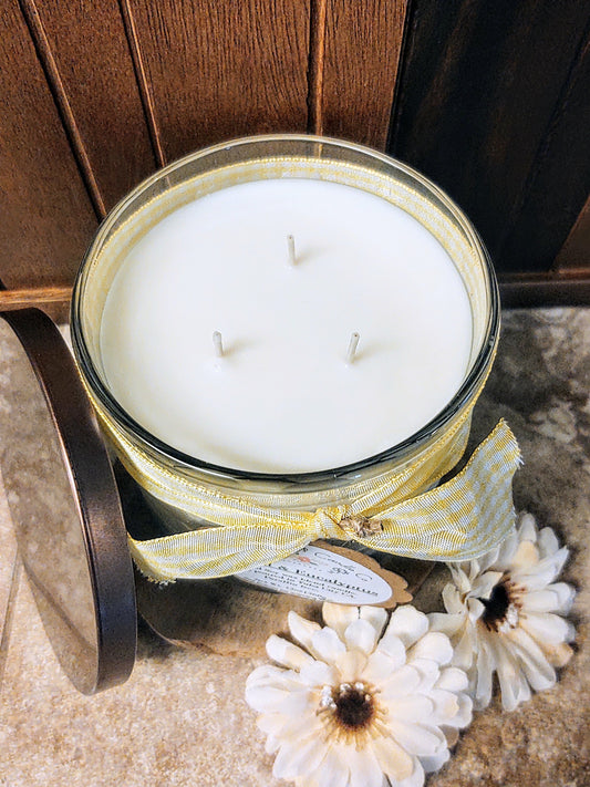 A large 3-wick natural soy wax candle in a glass jar with a yellow ribbon on a wooden surface with flowers. Eucalyptus scented candle.